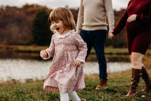 Child in floral pink dress and white tights runs joyfully outdoors near water with adults visible in background.