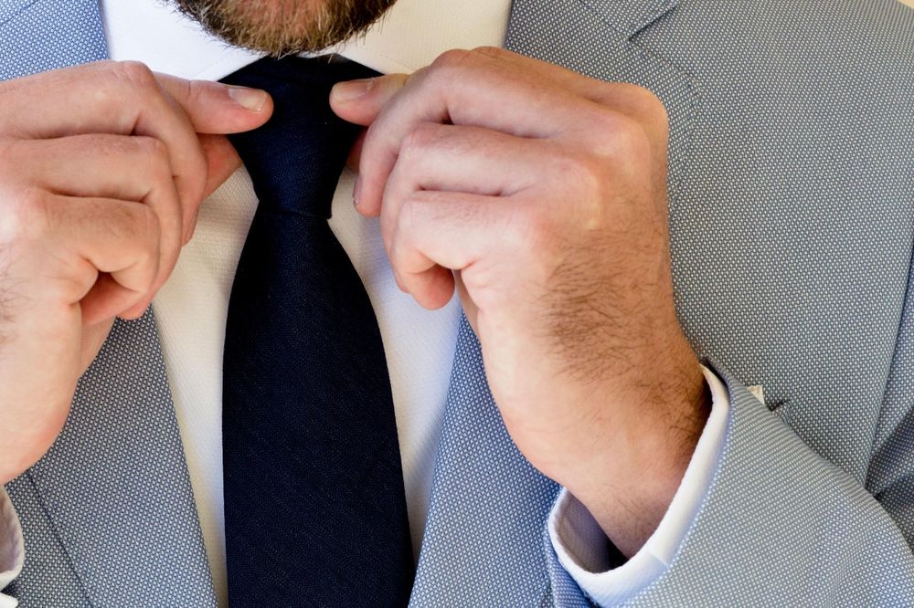 A close-up shot shows hands adjusting a blue necktie against a light-colored suit.