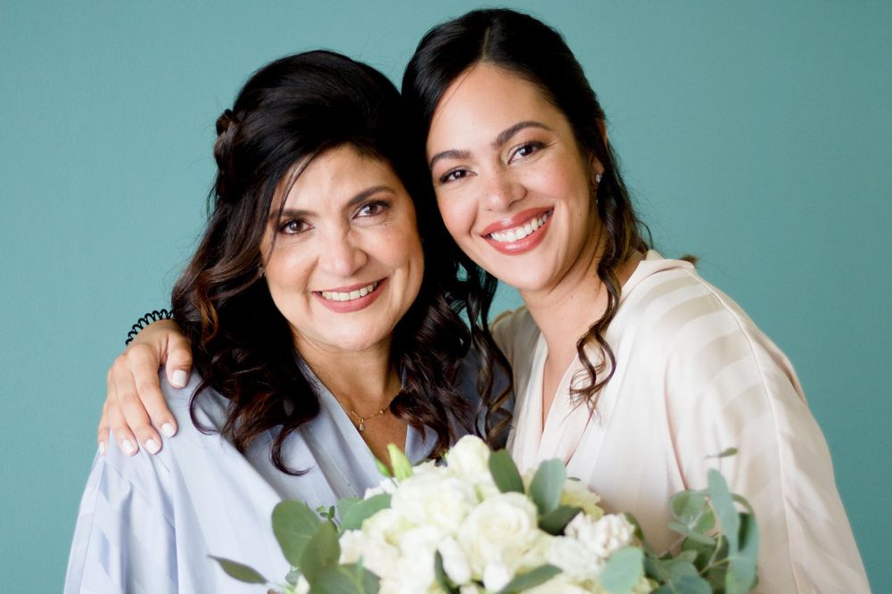 Two people in white tops pose together against a turquoise background with white flowers.