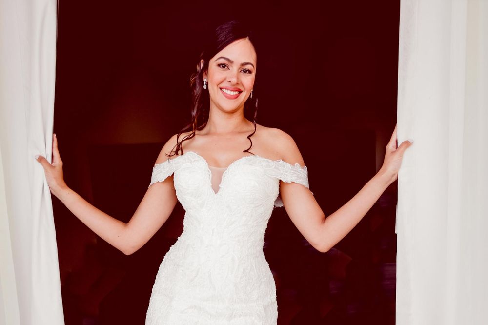 A bride in an elegant off-shoulder white lace wedding dress poses between white curtains against a dark red background.