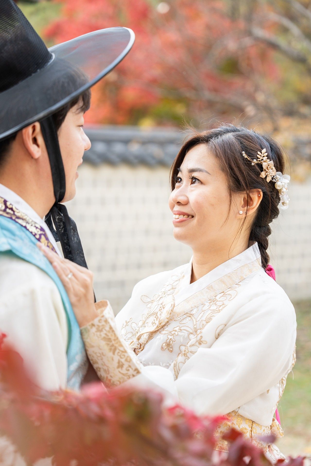 Traditional Korean couple in hanbok attire share joyful moments together during autumn with red maple leaves in background.