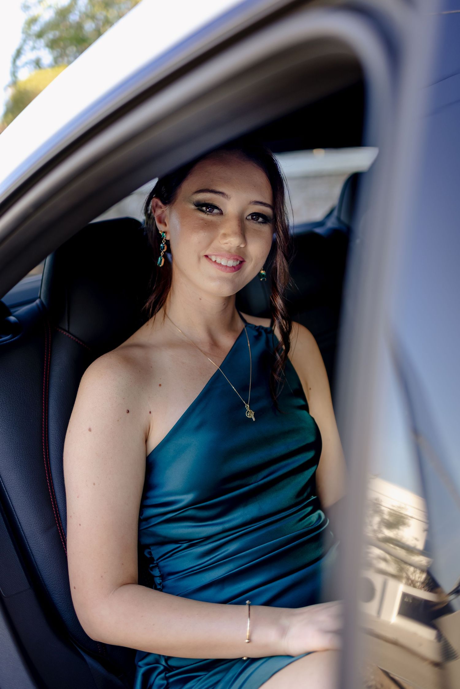 A woman in a teal one-shoulder dress poses elegantly inside a car.