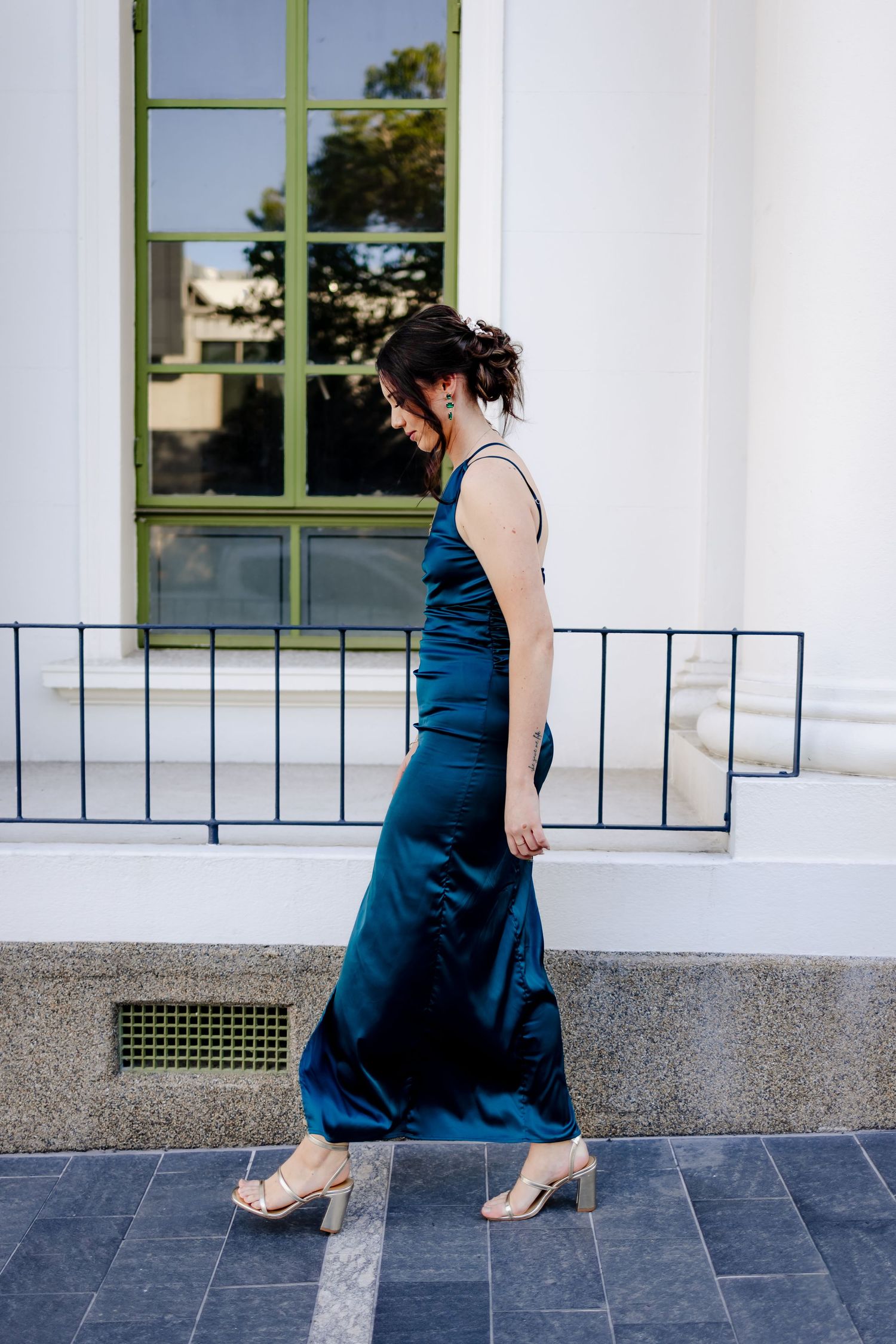 A figure in a flowing navy blue evening gown poses against a white columned building with green-framed windows.