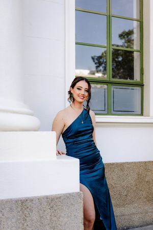 A blue evening gown is elegantly displayed against a white building exterior with green-trimmed windows.