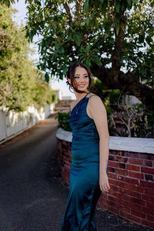 Woman in teal evening gown stands beside brick wall on garden pathway at dusk.