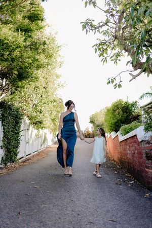 Mother and daughter walk hand in hand down a tree-lined path on a sunny day.