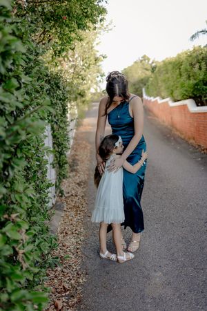 Child in white dress and woman in navy gown share tender moment on garden path.