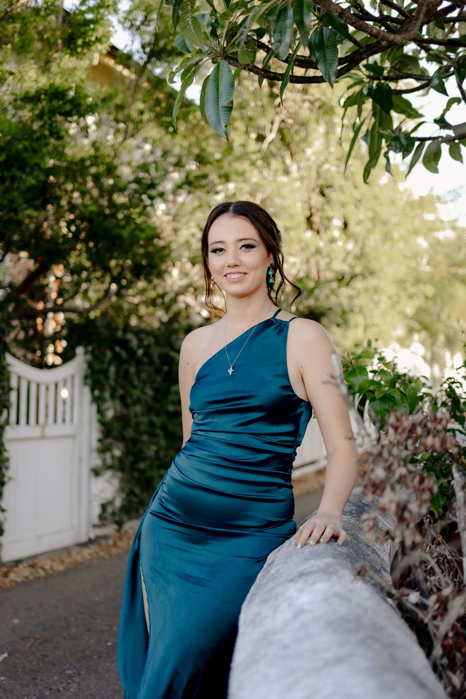 A woman in a deep blue formal dress poses outdoors among green foliage.
