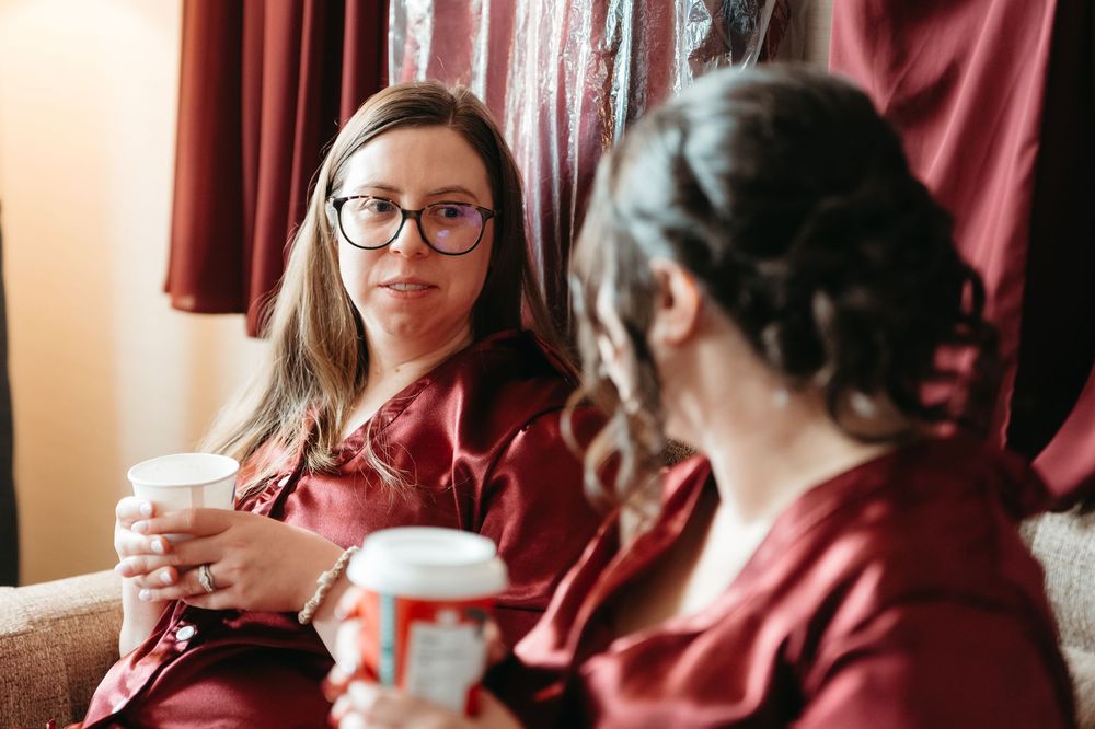 A person in glasses holds a white coffee cup while sitting near red curtains in a warmly lit room.