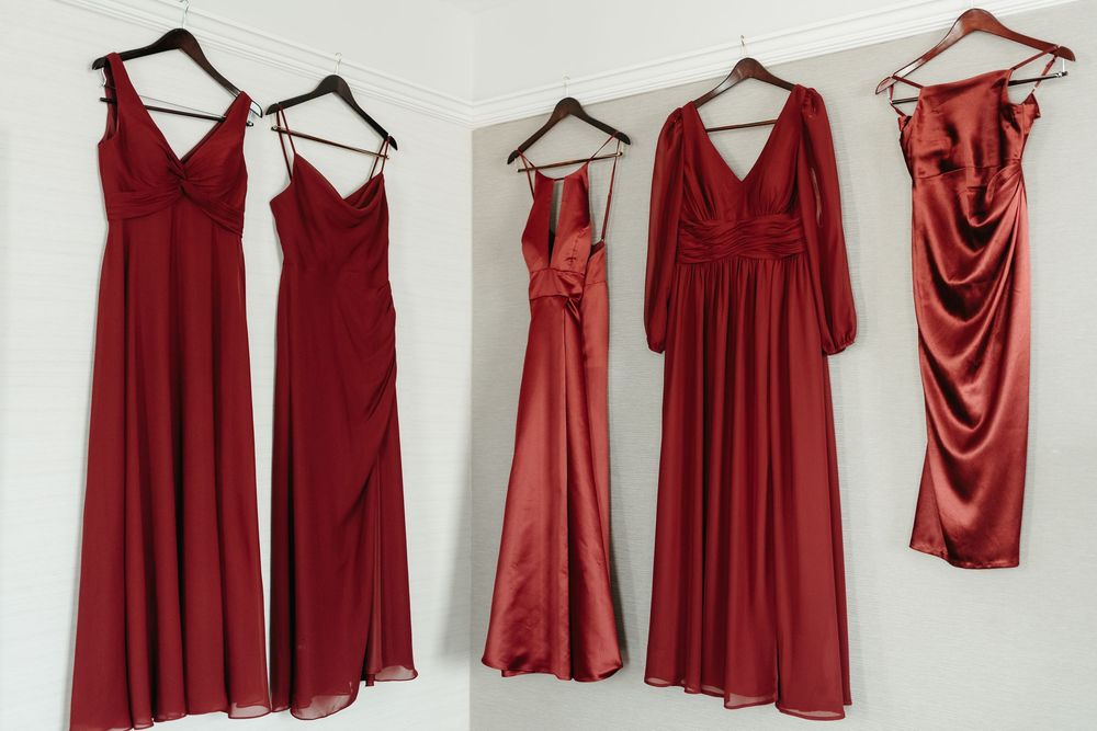 A collection of red and burgundy bridesmaid dresses hanging on white hangers against a light colored wall.