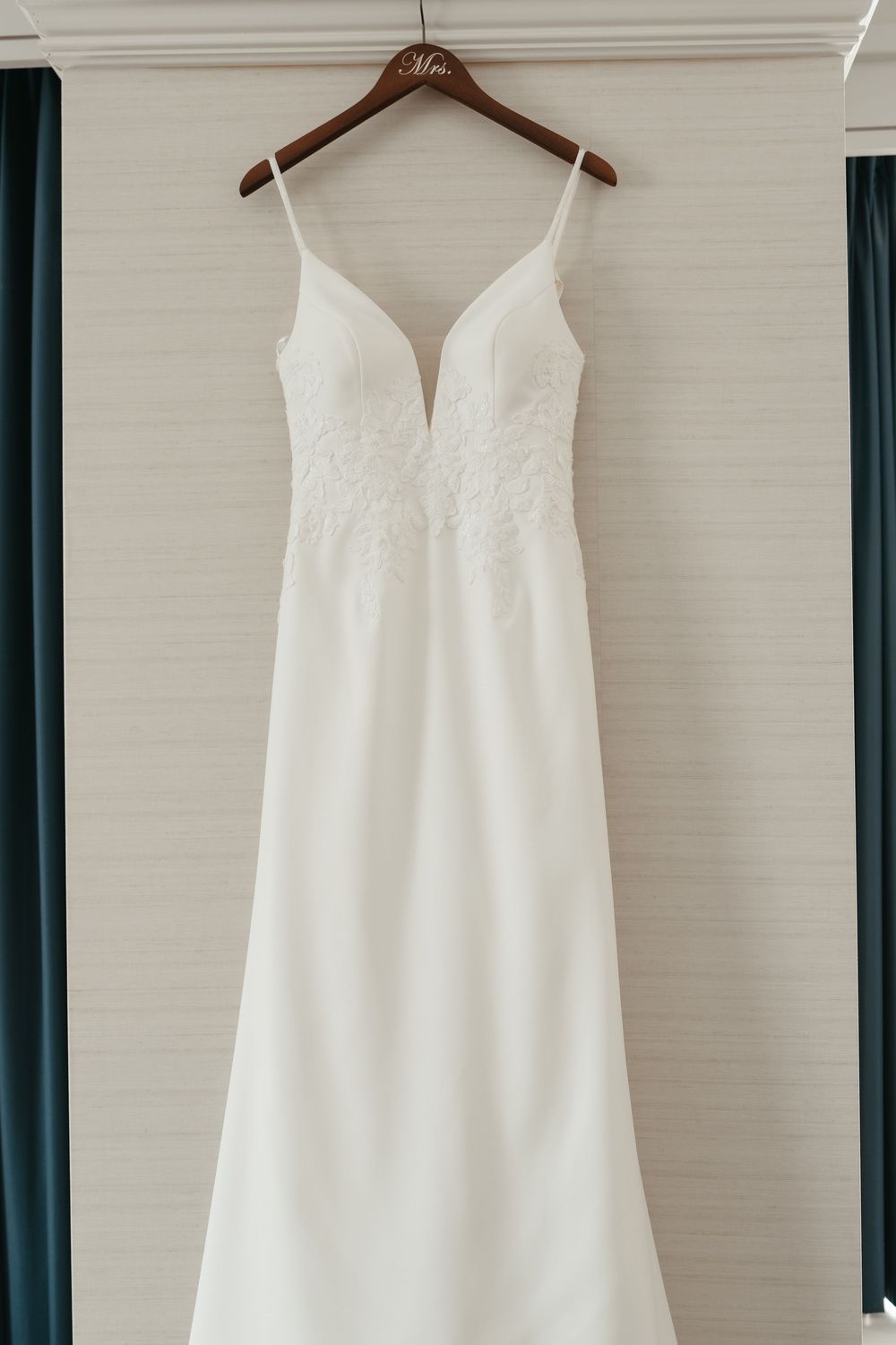 Three elegant white wedding dresses hanging on wooden hangers against a light colored wall with dark curtains.