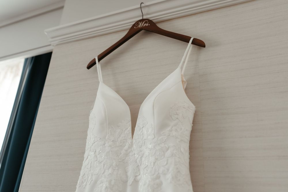 An elegant white lace wedding dress with plunging neckline hangs on wooden hanger against light colored wall.