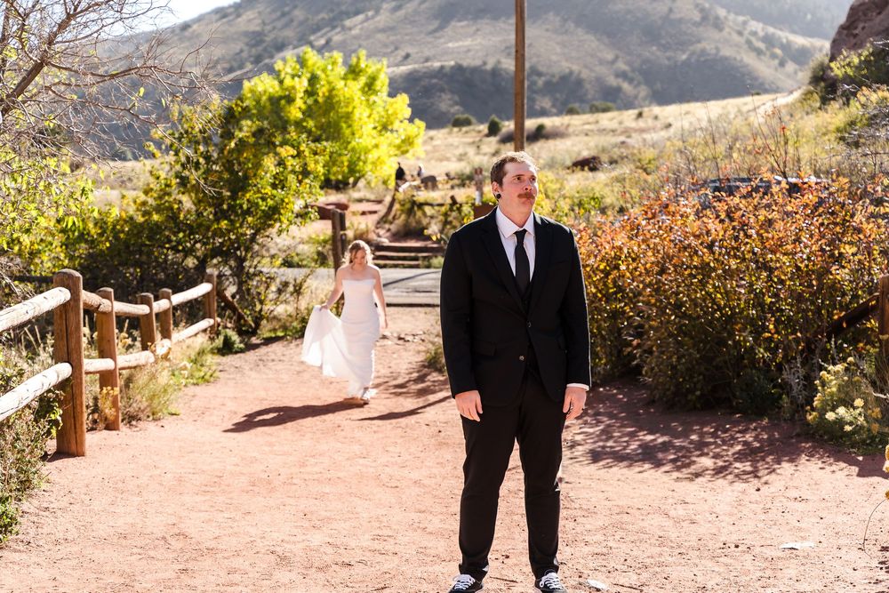 A couple in wedding attire walks hand in hand along a scenic mountain trail during a sunny outdoor ceremony.
