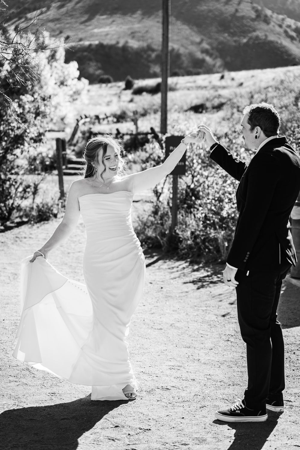 A couple shares a joyful dance moment on a dirt path with mountains in the background in black and white.