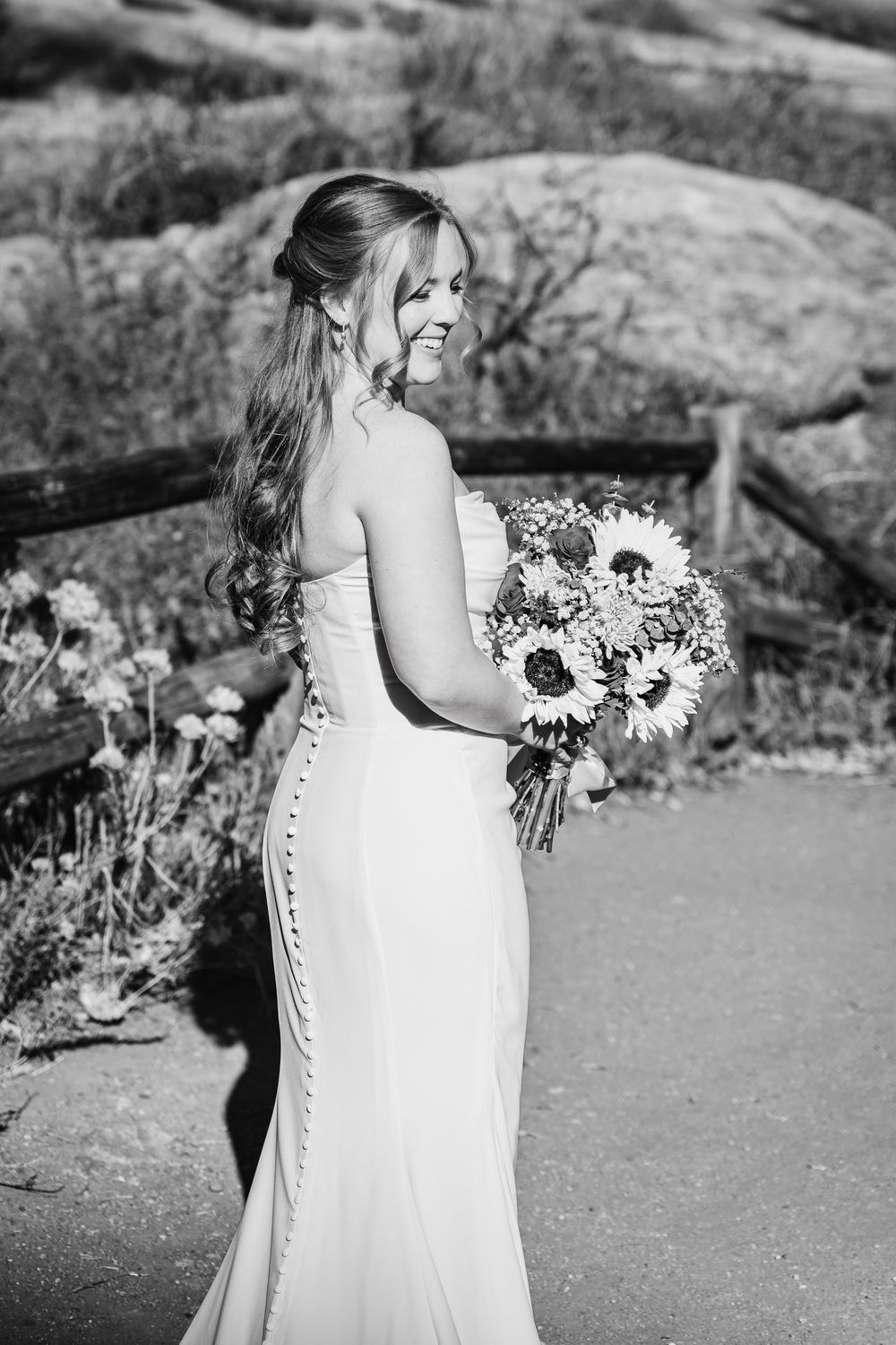 Black and white wedding portrait showing a bride in a button-back dress holding sunflowers in a natural outdoor setting.