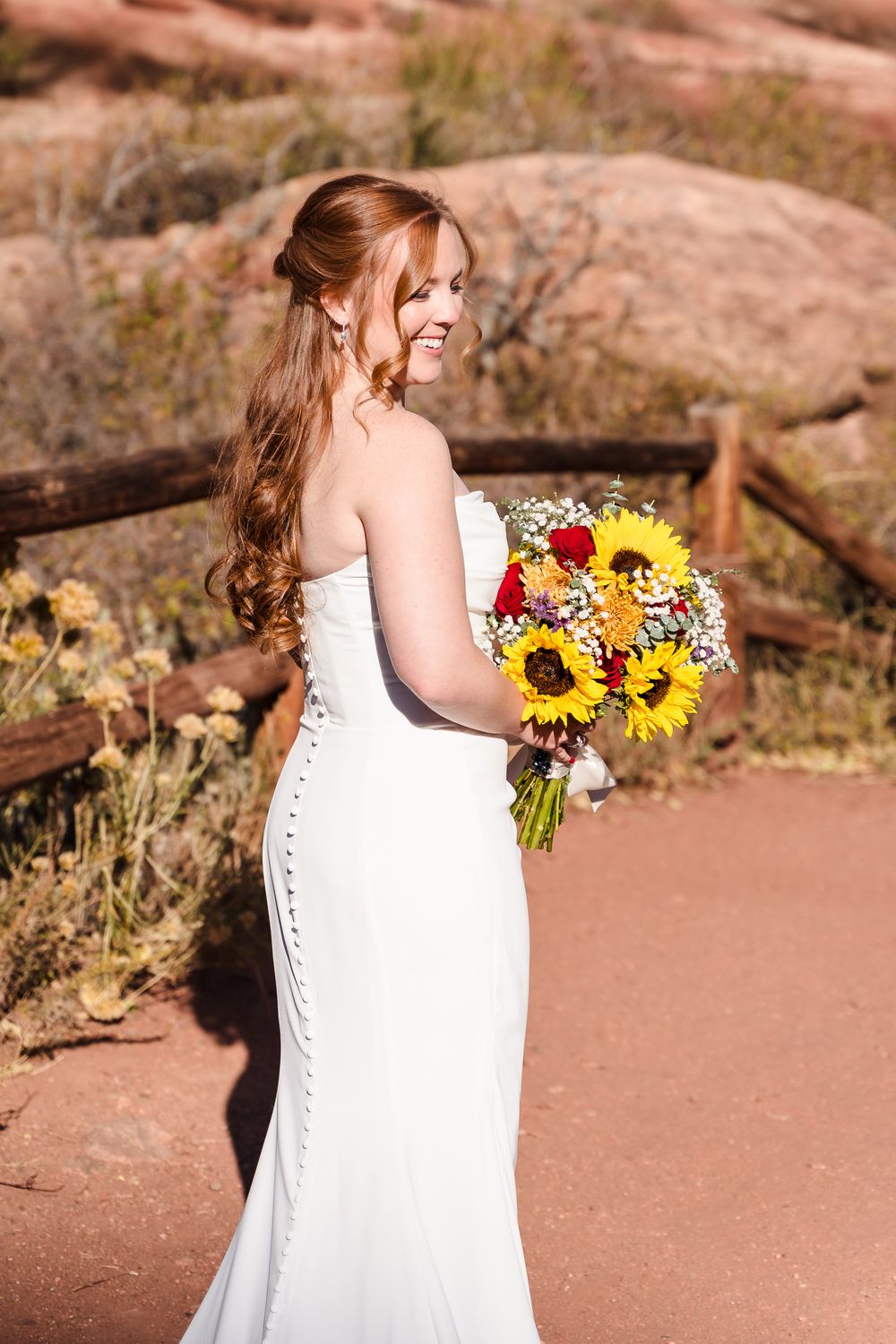 A bride in a fitted white wedding dress holds a vibrant bouquet of sunflowers and red flowers against a desert landscape.