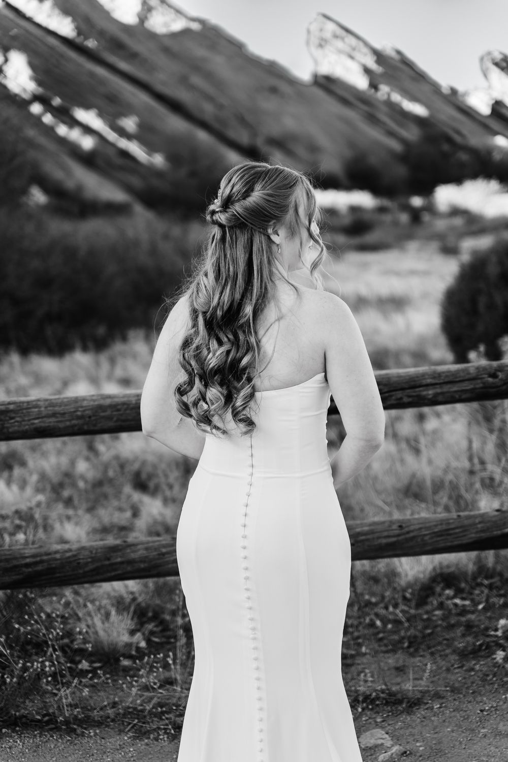 A bride in a classic white wedding dress with button details stands by a rustic wooden fence at sunset.