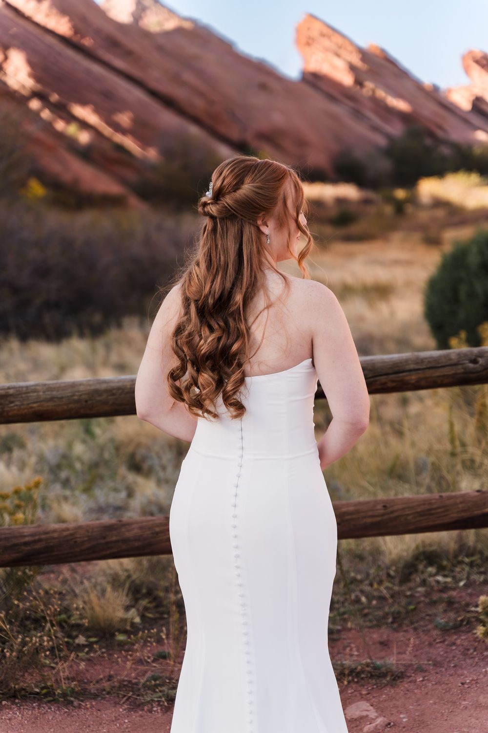 A bride in a white dress with button details stands at a wooden fence against red rock formations at sunset.