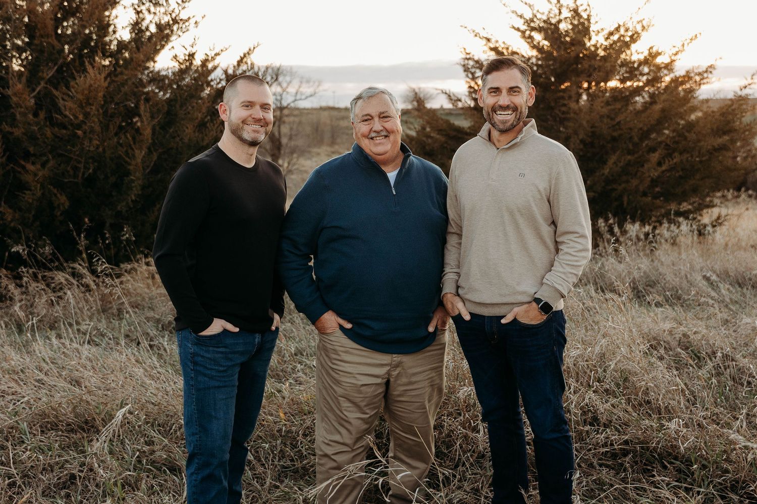 Three people stand together outdoors in a field at sunset with shrubs and tall grass in the background.