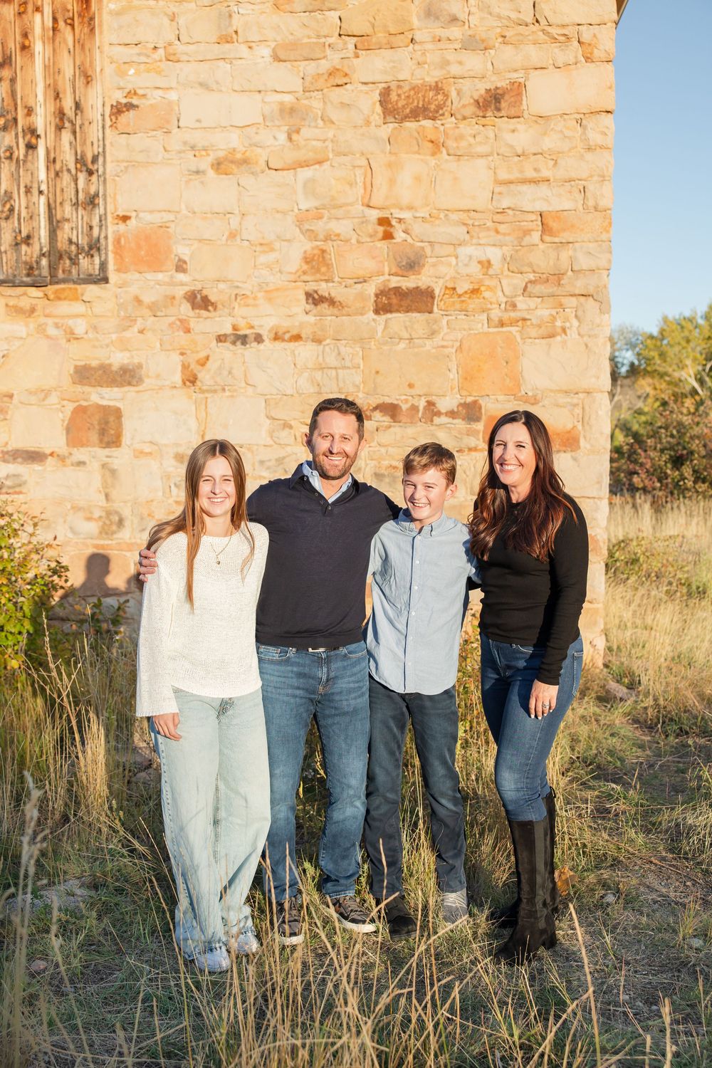 A group poses together against a rustic stone wall during golden hour in a field of tall grass.