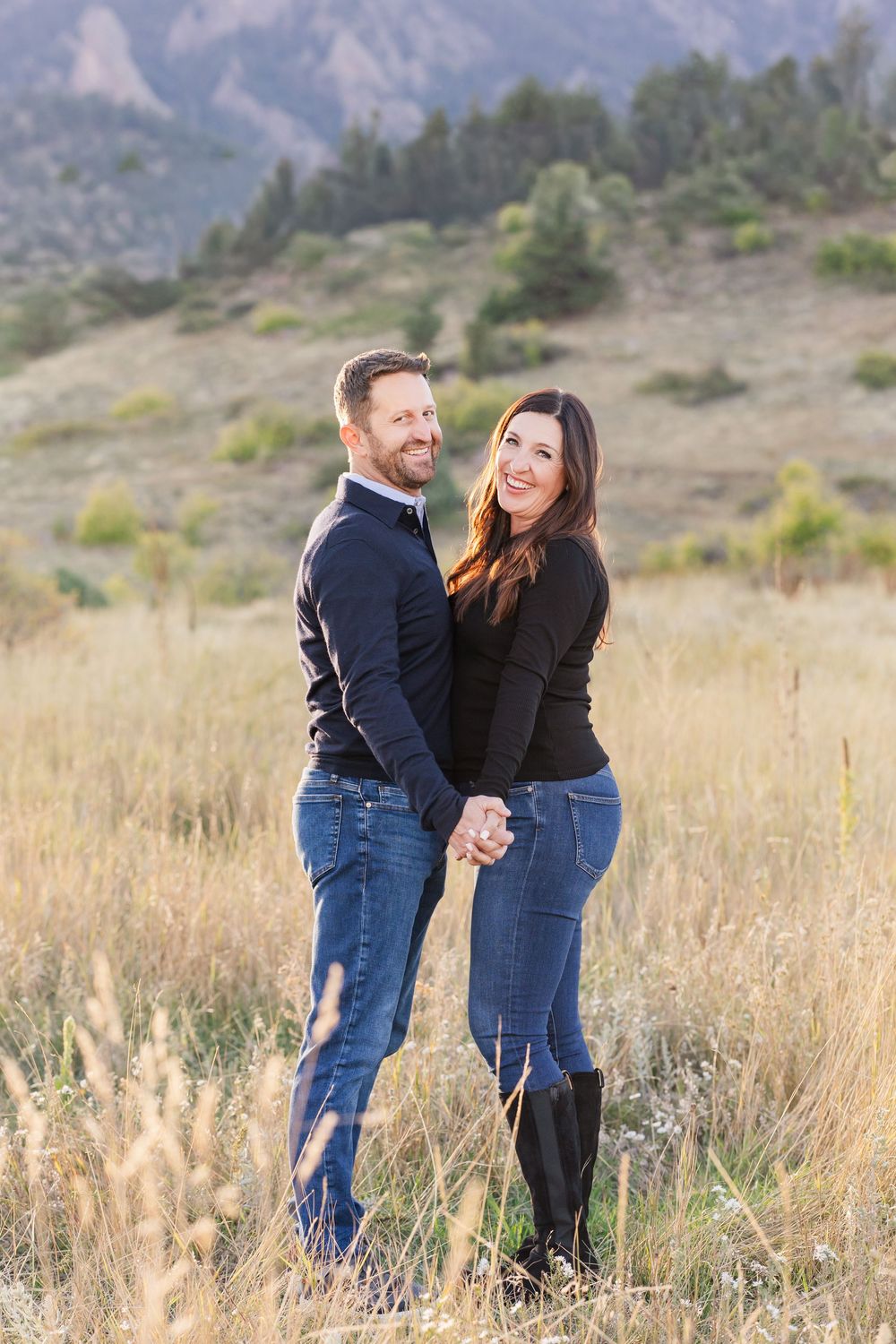 A couple in casual black tops and jeans embrace while standing in a golden field with mountains in the background.