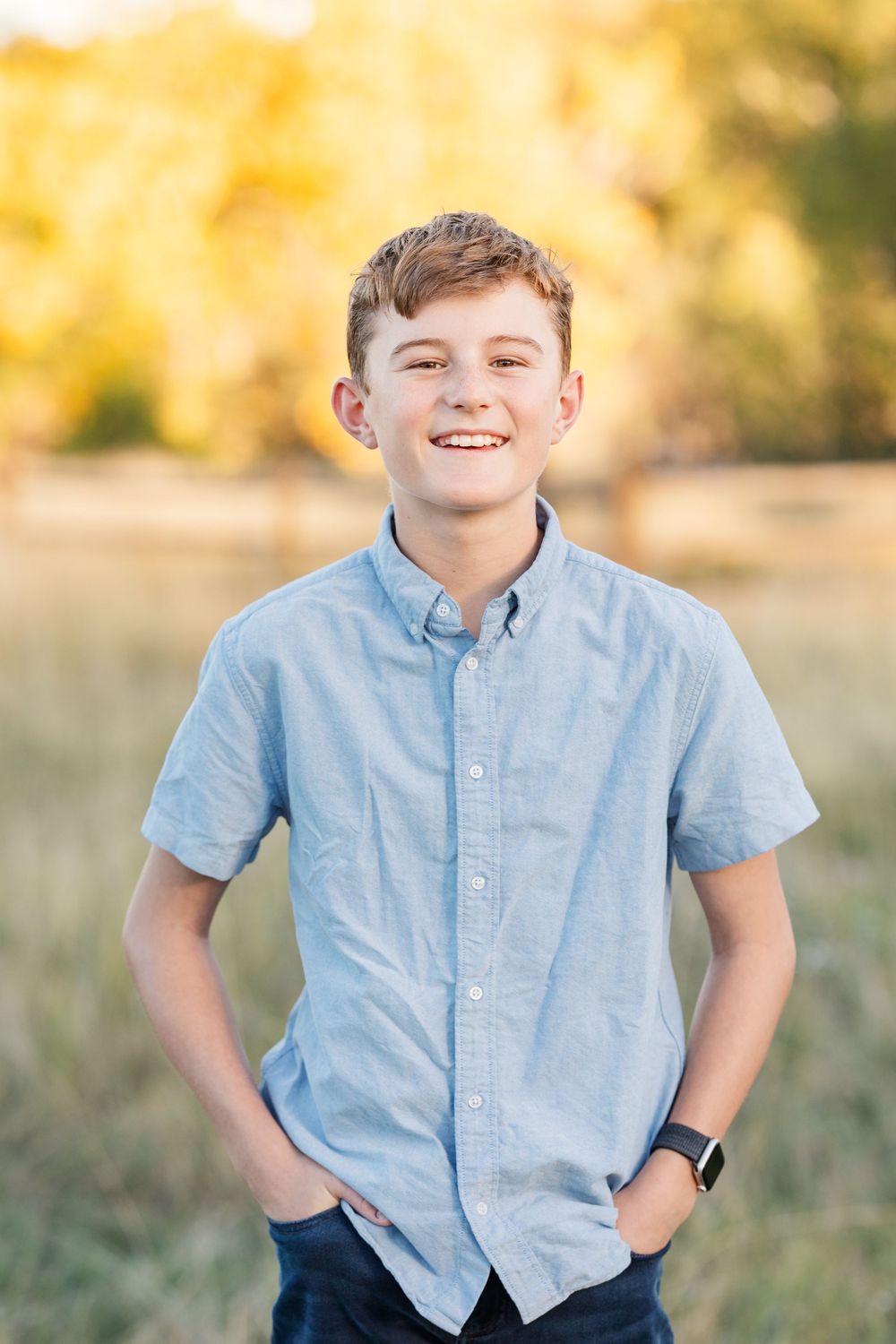 Young boy in light blue shirt smiling outdoors in golden sunlight.
