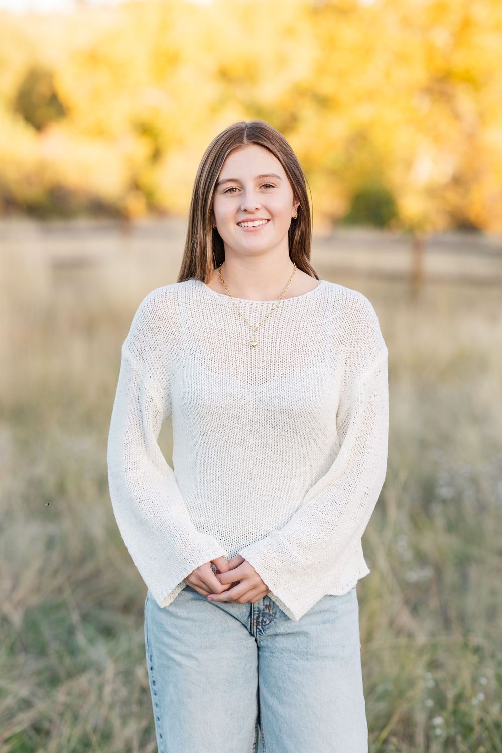 Person in white sweater and light jeans poses outdoors during a fall photoshoot with golden foliage.