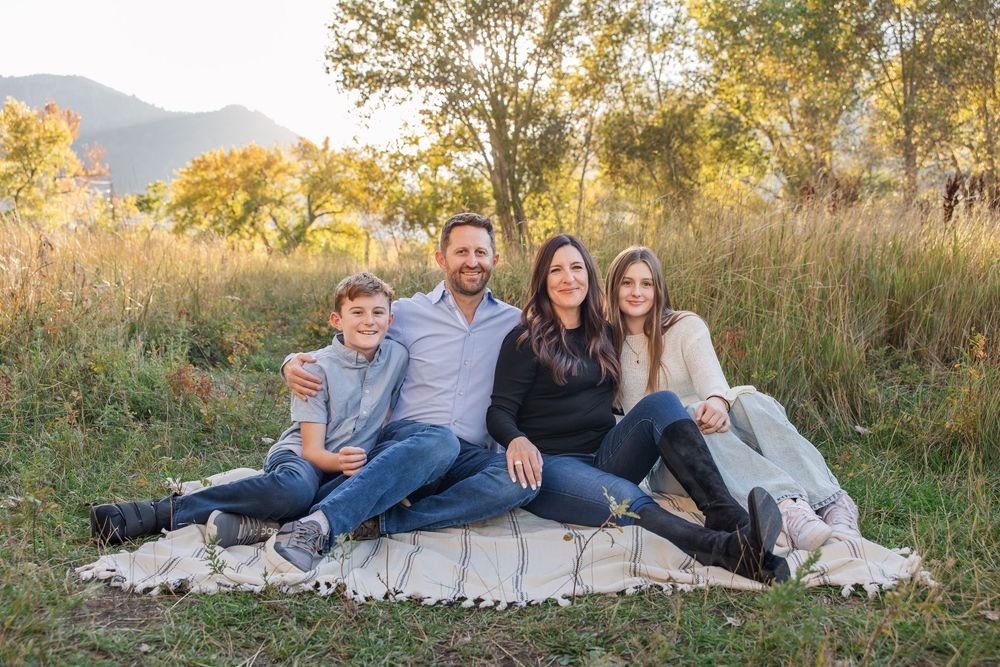 Family lounging together on blanket in grassy field with mountains and autumn trees in background at sunset.