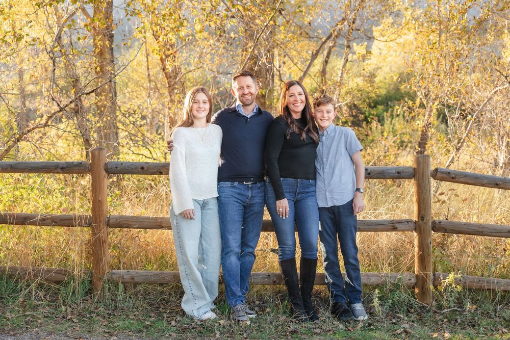 A group poses together outdoors against a rustic wooden fence with autumn foliage in the background.