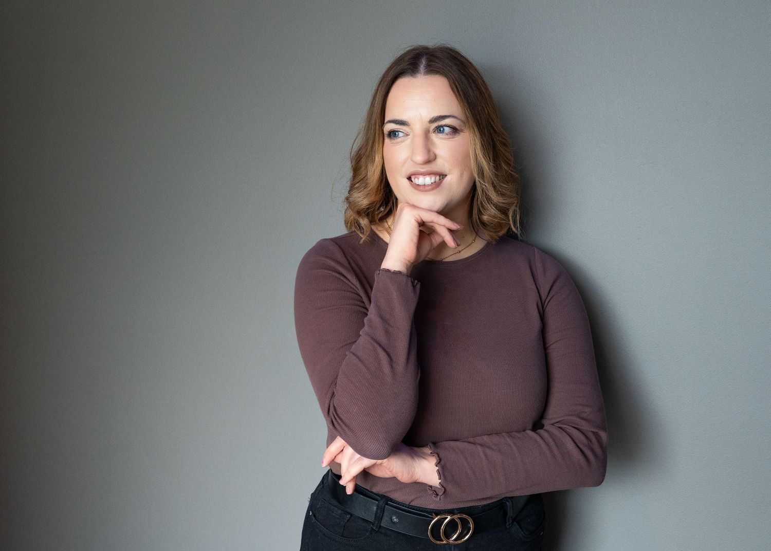 Person in brown sweater and jeans poses confidently against grey backdrop in professional photo series. Headshots in Glasgow.