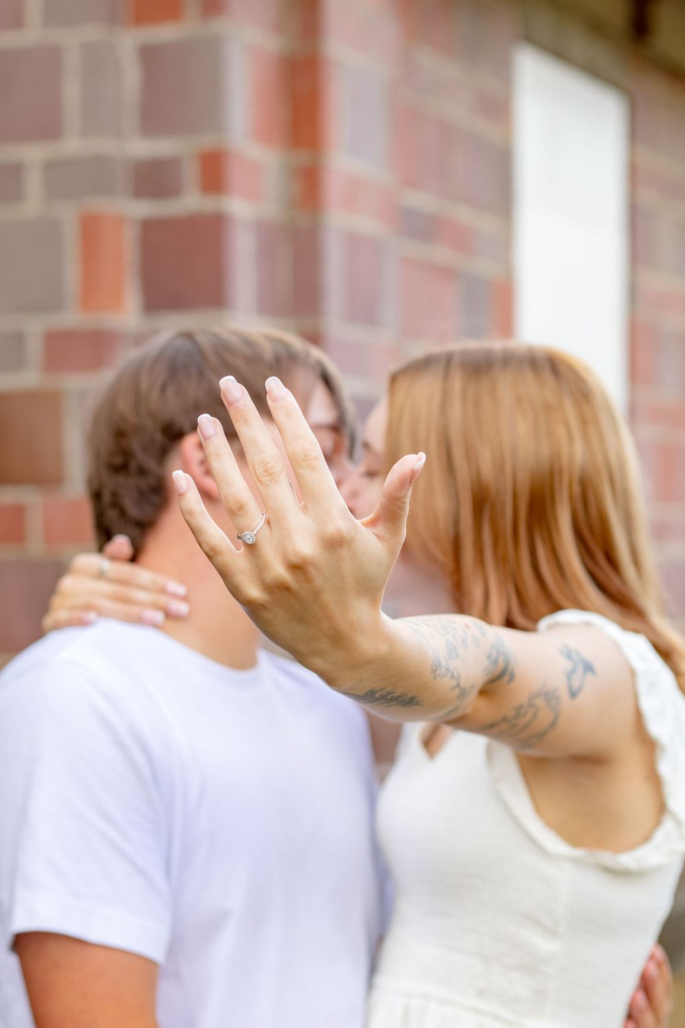 An engaged couple kisses while the woman displays her diamond engagement ring and tattooed arm toward the camera.