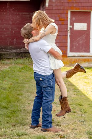 A couple shares a romantic kiss as the man lifts the woman in a white dress and cowboy boots near a red barn.