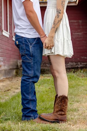 A couple holds hands near a red barn, the woman wearing a white dress and brown cowboy boots with visible tattoos.