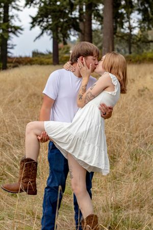 A man lifts and kisses his partner in a white dress and cowboy boots in a golden field surrounded by tall pine trees.