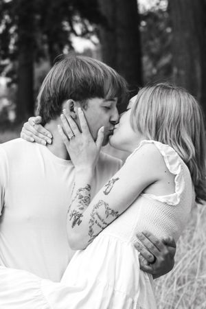 A black and white romantic portrait of an engaged couple sharing a kiss, the woman displaying her ring and tattoos.