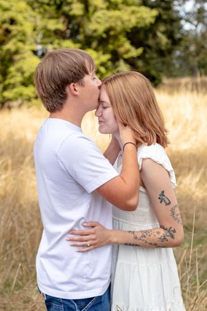 A young couple shares a tender forehead kiss in a golden field, the woman showing her floral arm tattoos.