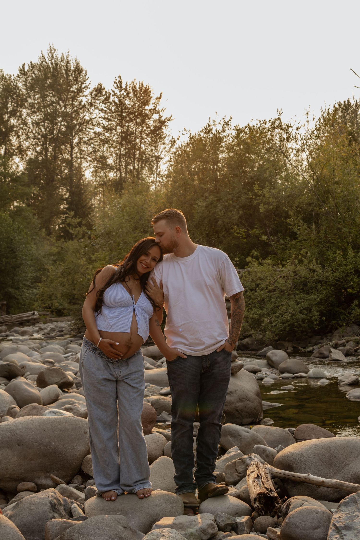 Couple standing on rocky little mashel riverbank at golden hour, woman leaning on man as he kisses her head near lush green trees.