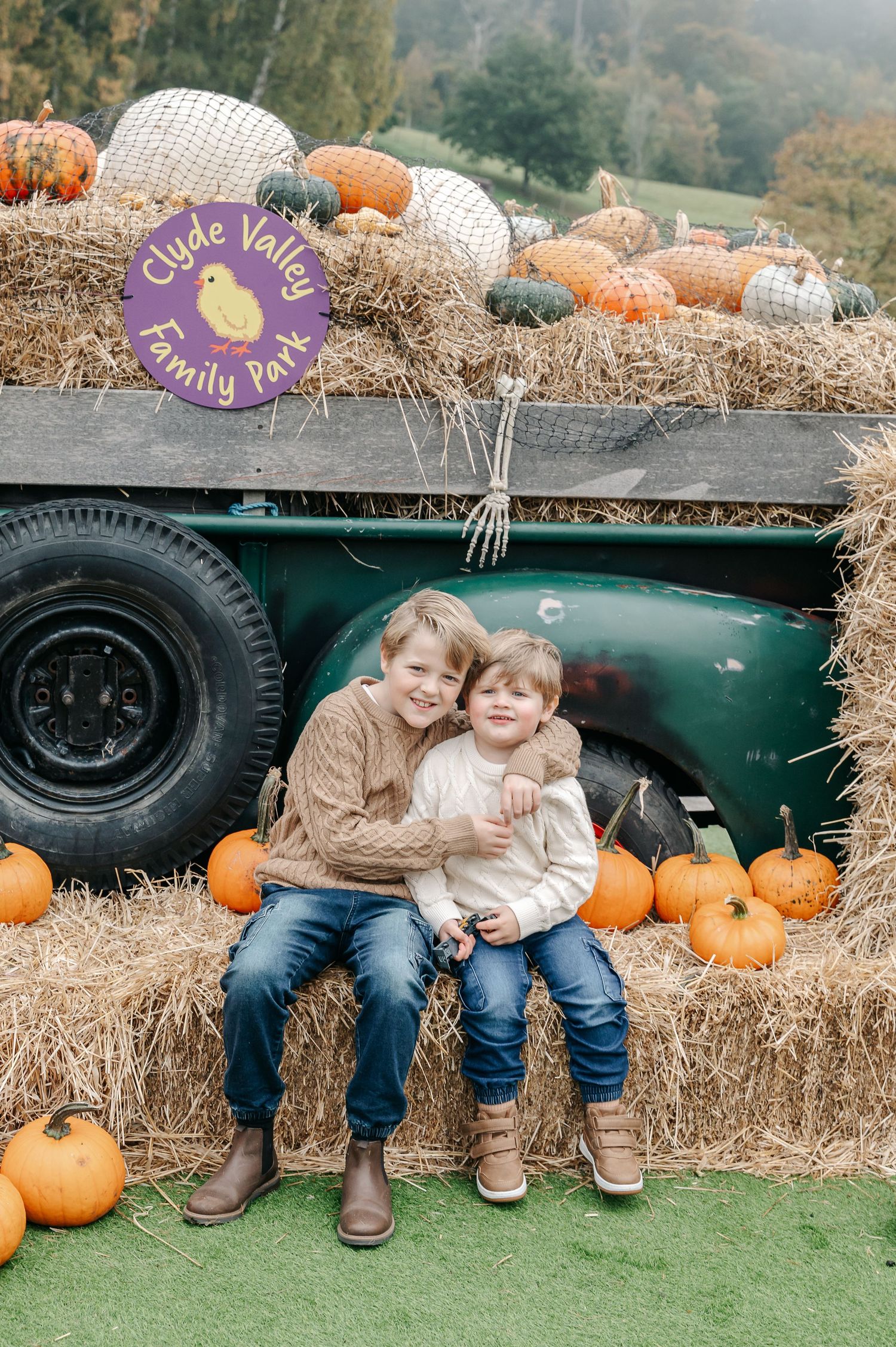 Children sitting on hay bales next to pumpkins and a vintage green truck in an autumn setting. Branding for Clyde Valley Family Park.