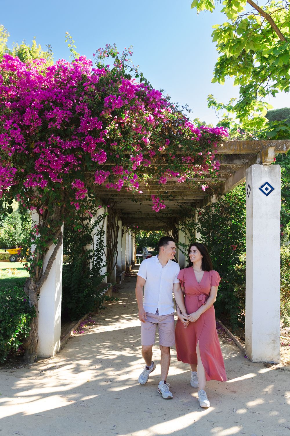A couple strolls under a beautiful pergola covered in vibrant pink bougainvillea flowers in a garden setting.