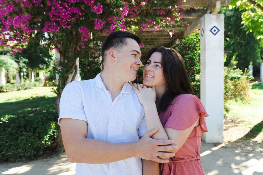 A couple shares a romantic moment under a blooming pink bougainvillea pergola in a garden setting.