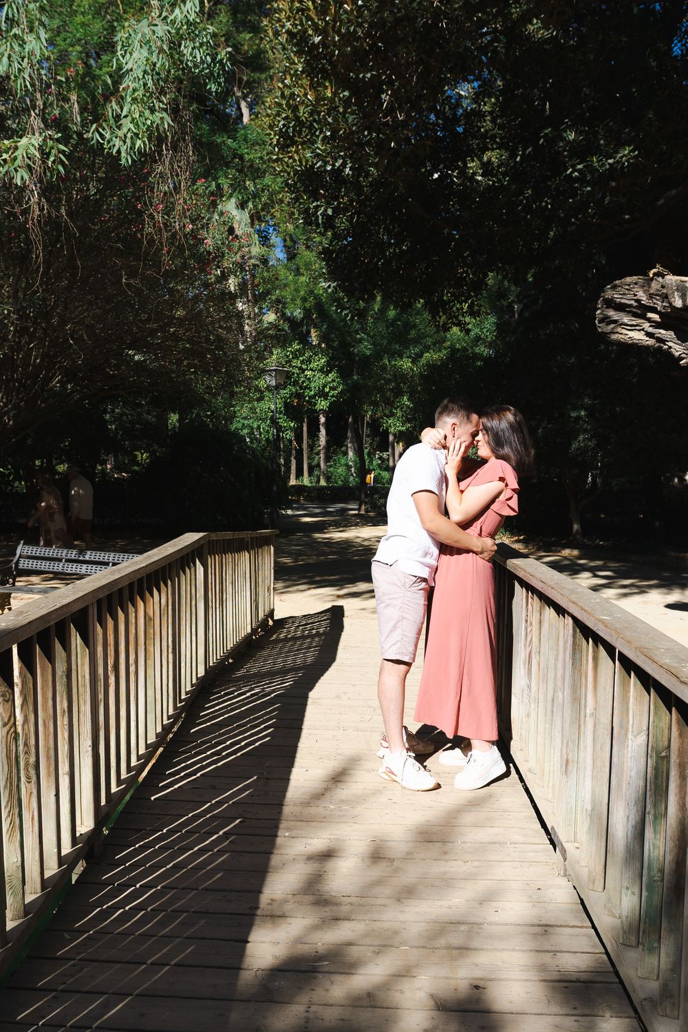 A couple in formal attire share a romantic moment on a wooden boardwalk surrounded by lush greenery.