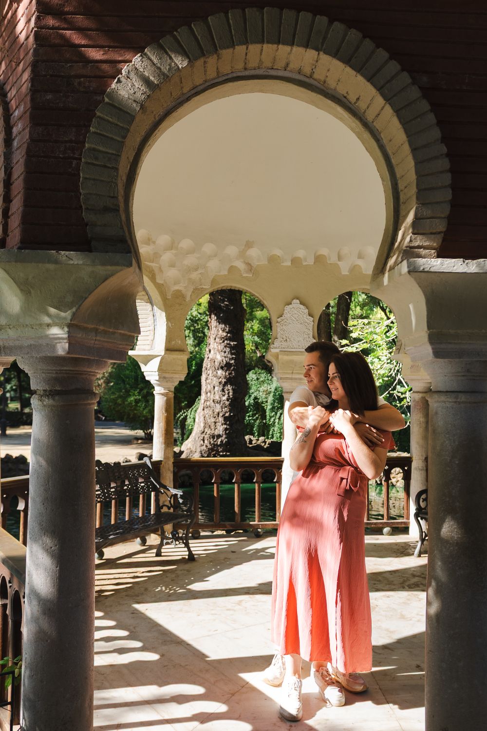 Two people in elegant attire stand beneath ornate stone archways with sunlight streaming through.