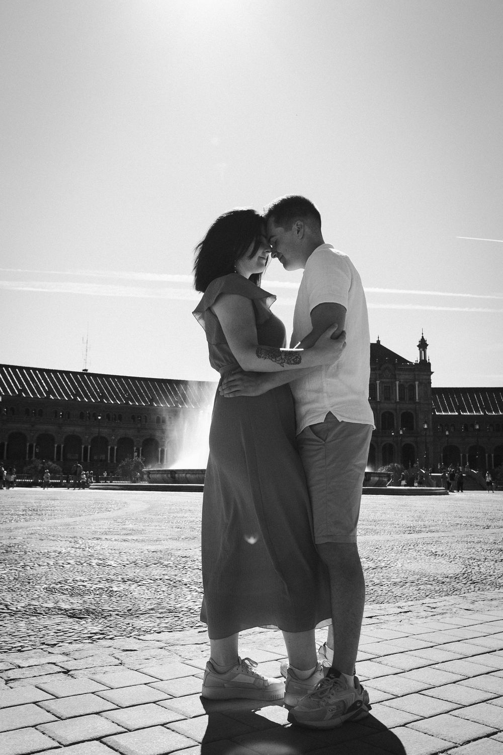 A couple shares an intimate moment in silhouette against a bright sky with historic architecture in the background.