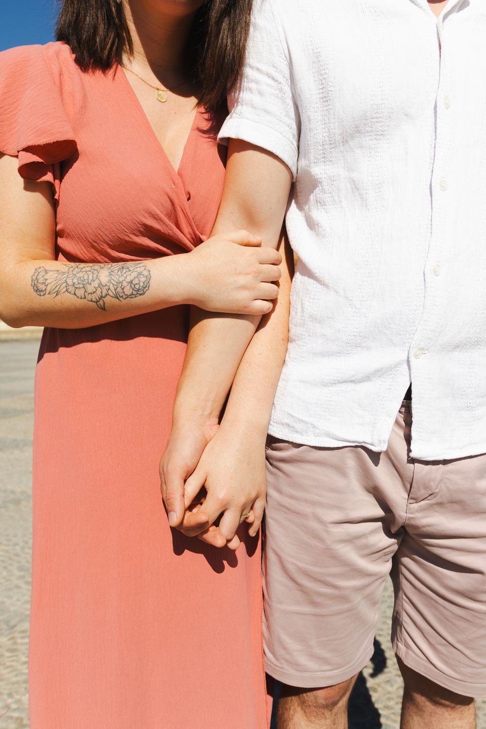 Close-up of a couple holding hands, showing a coral dress, white shirt, and visible tattoo detail.