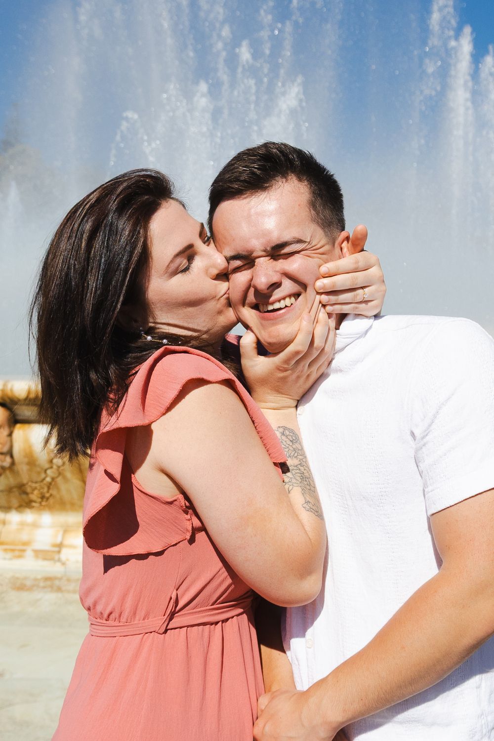 A couple shares a playful moment in front of a fountain, with one person in a coral dress and another in white.
