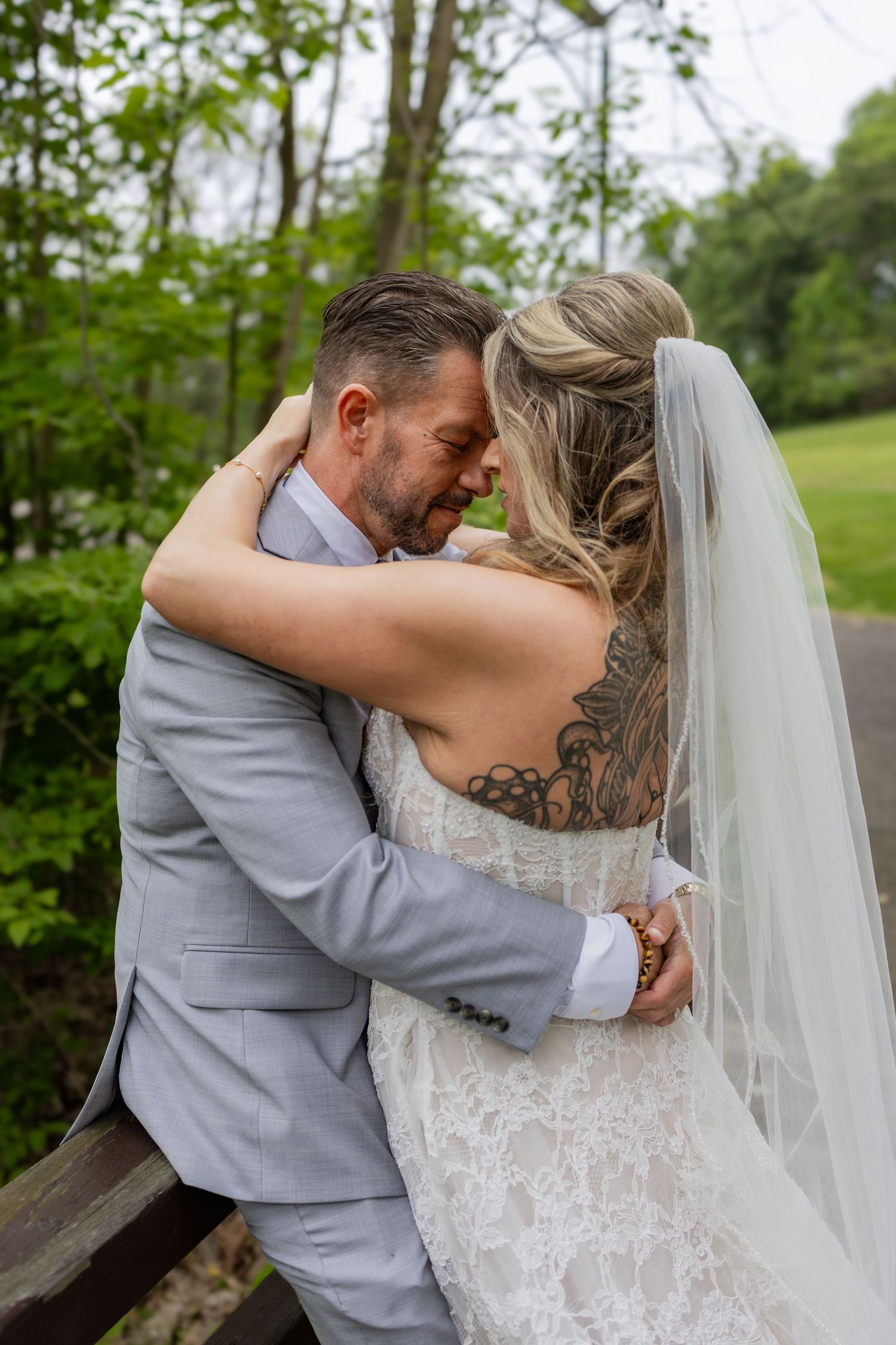 Bride and groom share intimate embrace on bridge, bride wearing lace gown with veil and back tattoo.