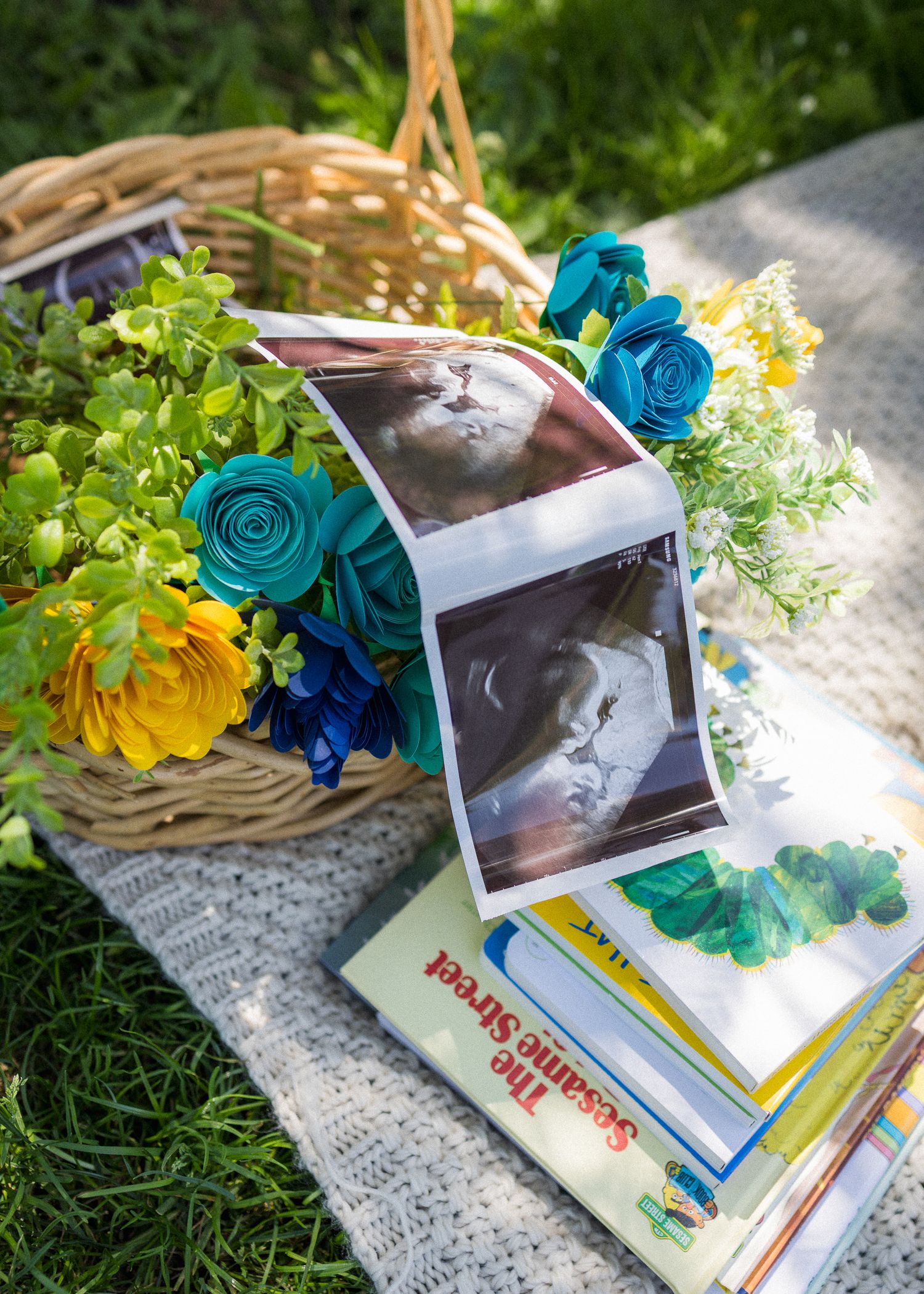 Floral arrangement with teal and yellow roses beside Sesame Street books and ultrasound photos in a wicker basket outdoors.