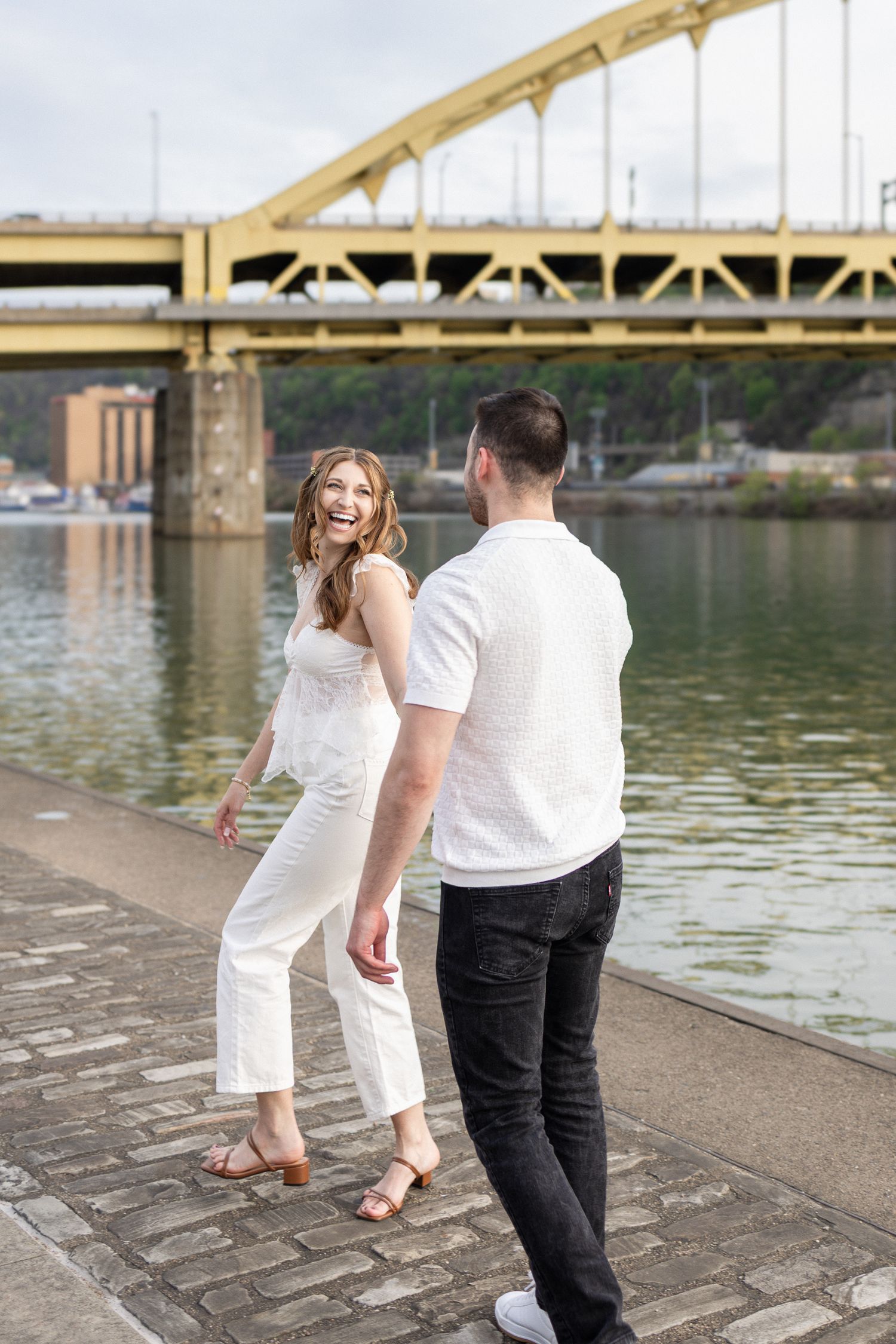 Couple holding hands by a river with a yellow bridge, woman in white outfit laughing joyfully on a cobblestone walkway.