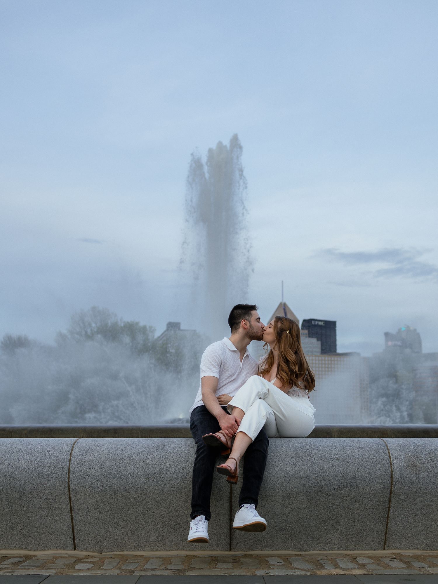 Couple sharing a romantic kiss on a stone wall with a misty fountain and city skyline in the background.
