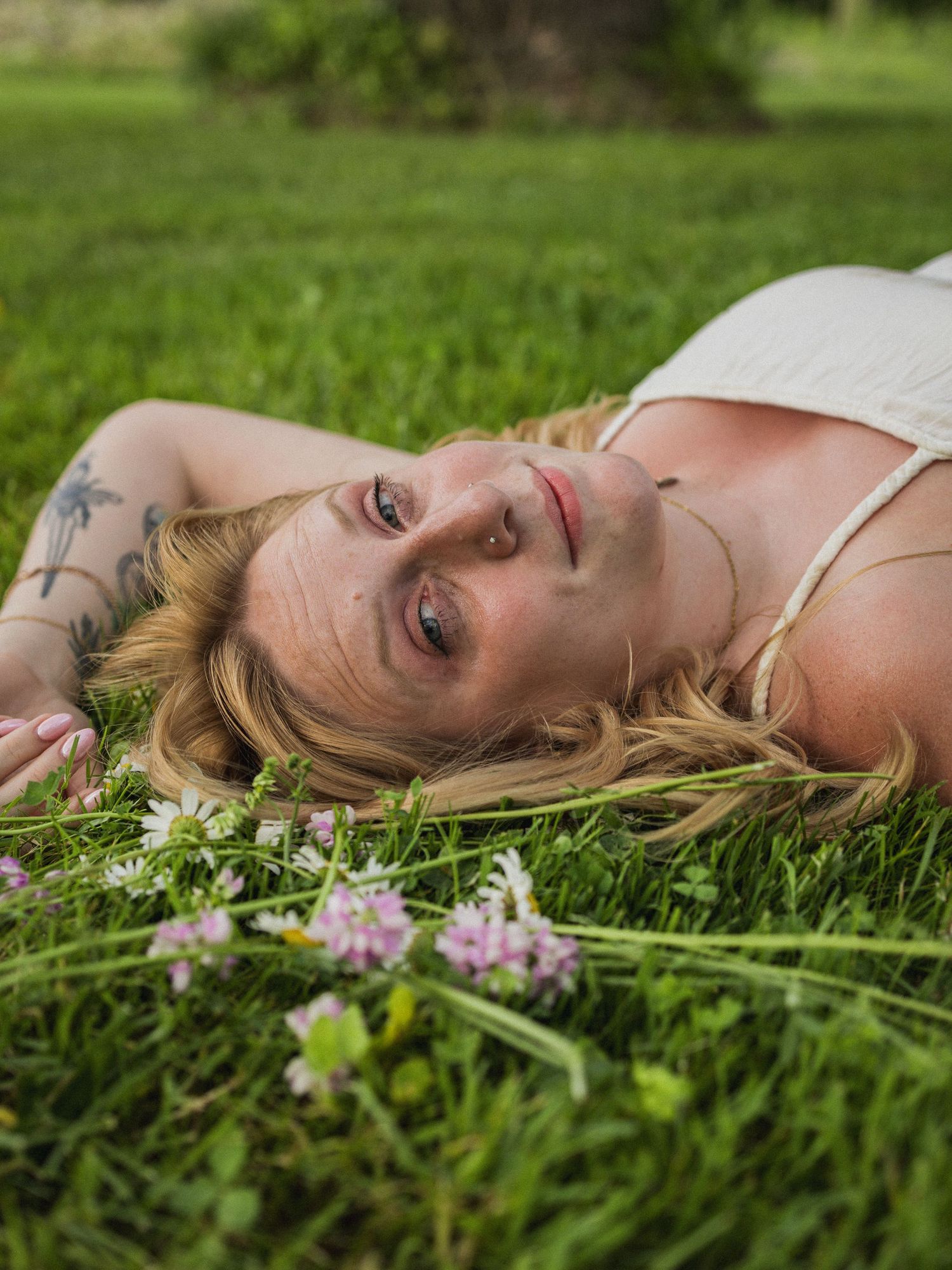 Blonde woman with tattoos lying in green grass surrounded by small wildflowers wearing a white top.